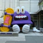 A large purple cartoon HiKR character inflatable sits outside a modern building, holding a yellow skateboard with pink graphics.