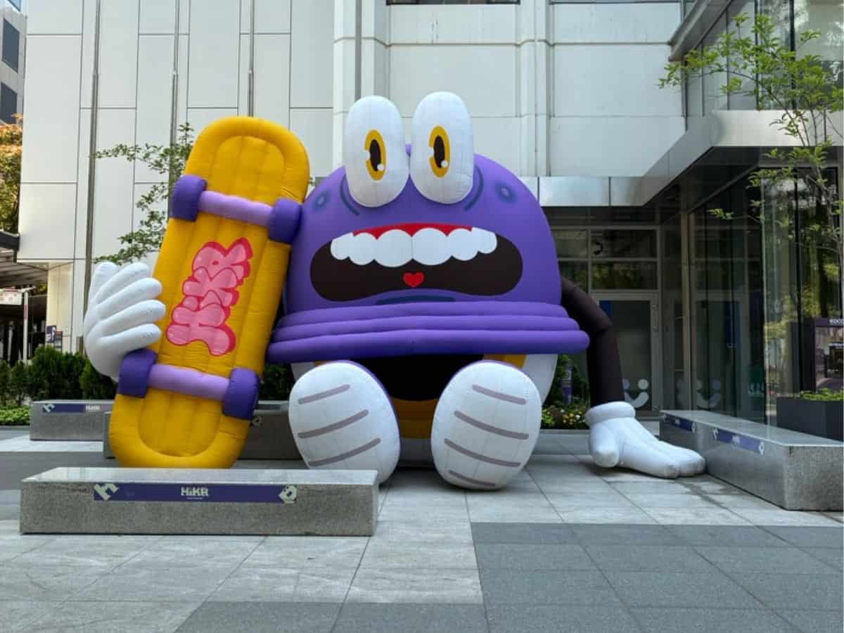 A large purple cartoon HiKR character inflatable sits outside a modern building, holding a yellow skateboard with pink graphics.