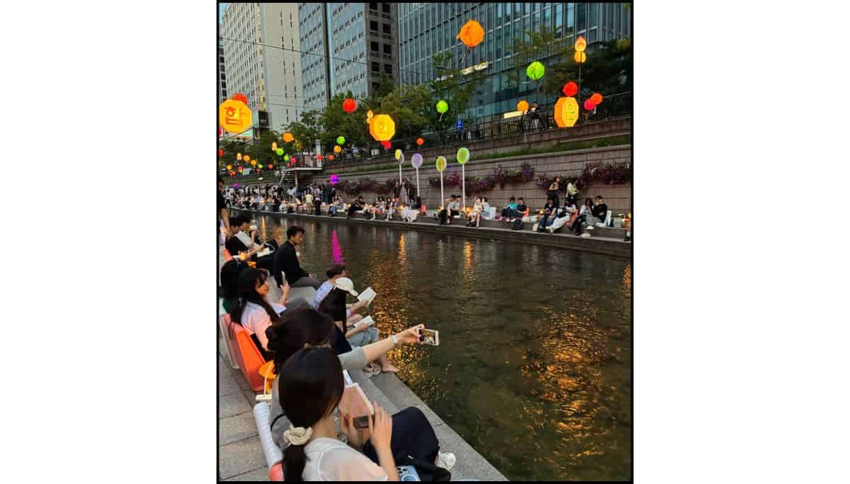 Busy evening scene at Cheonggyecheon Stream in Seoul, with people gathered by the water and lanterns overhead.