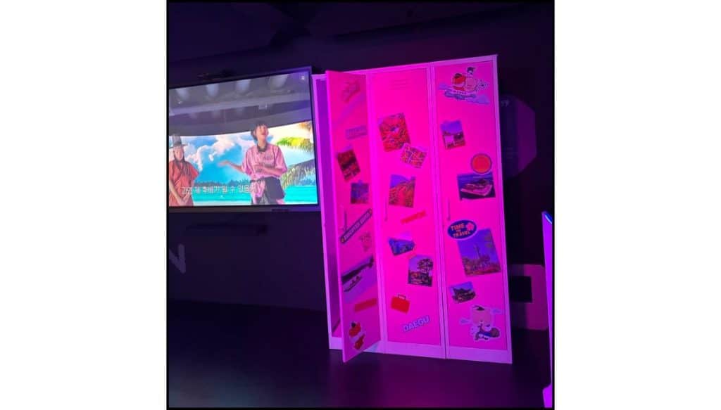 Pink-lit locker-style display covered with travel stickers, next to a screen playing a video inside HiKR Ground.