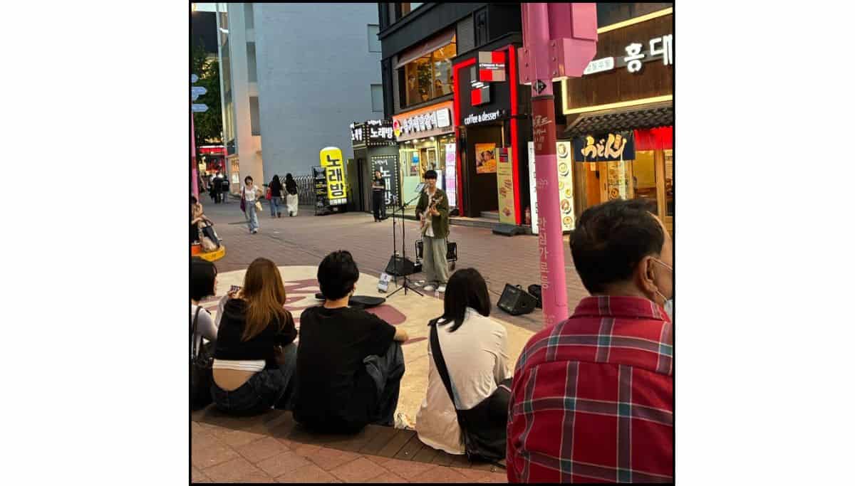 A street busker playing guitar to a crowd of people sitting on the ground in a busy Hongdae shopping street at night, surrounded by neon signs and Korean storefronts.