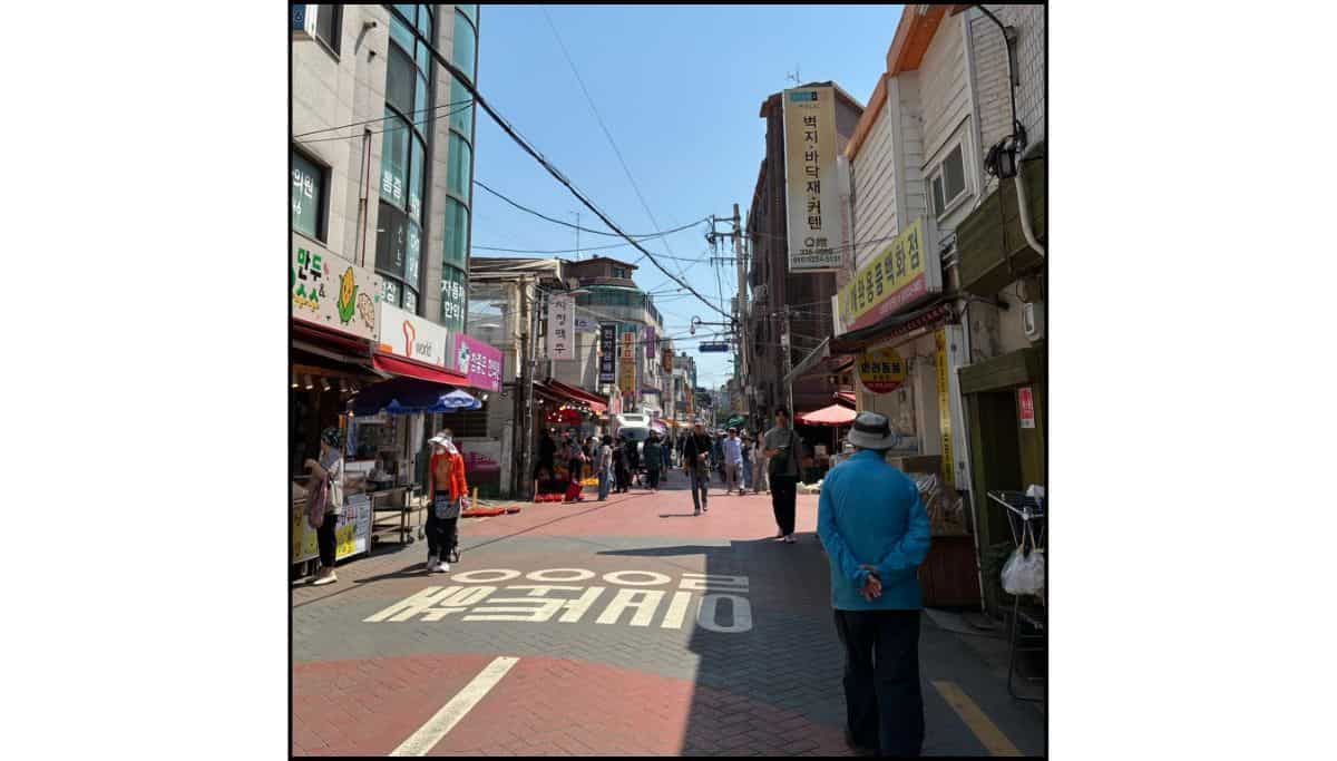 A busy pedestrian street lined with shops and street food stalls in a local Seoul neighborhood on a sunny day.