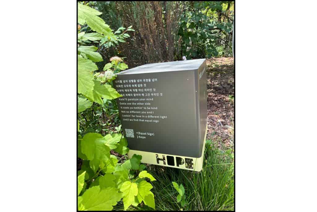 Metal box shaped sign displaying Korean and English lyrics from J-Hope’s “Equal Sign,” surrounded by green plants in J-Hope Garden at Seoul Forest.