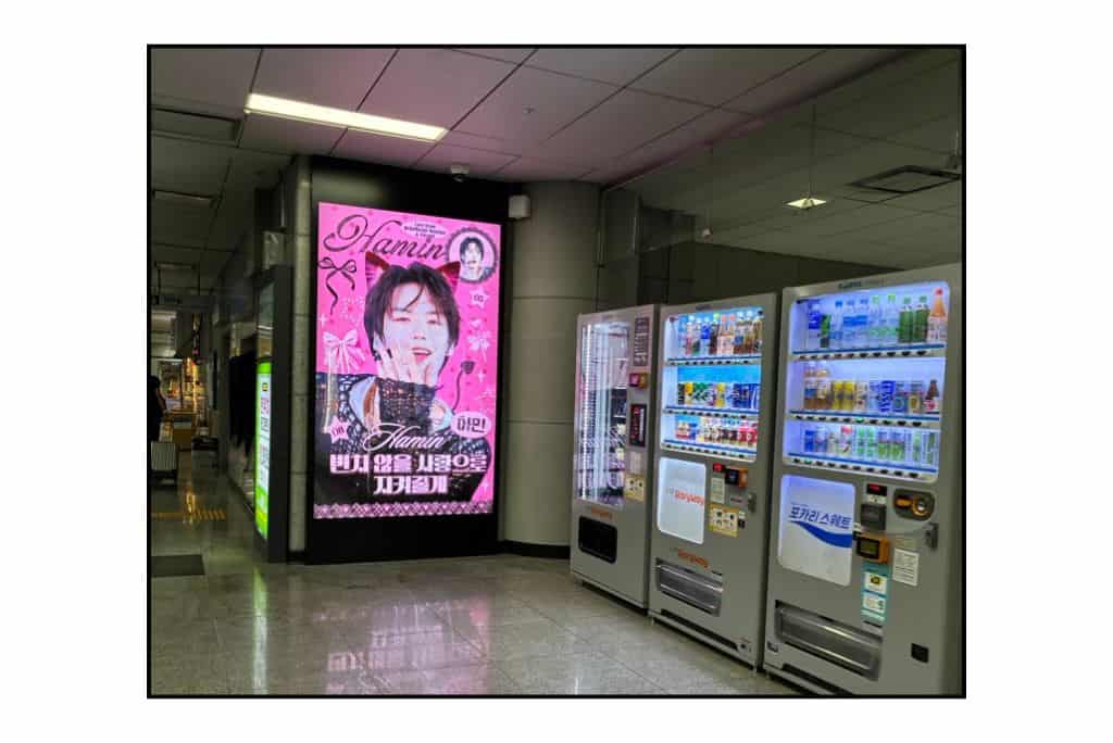 Pink digital birthday advertisement featuring SMTR25 trainee Hamin beside vending machines in a Seoul subway station.