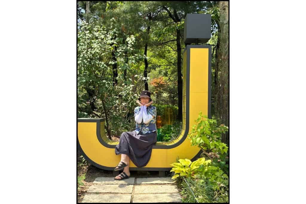 Woman posing on the base of a large yellow “J” sculpture surrounded by trees and plants in J-Hope Garden at Seoul Forest.